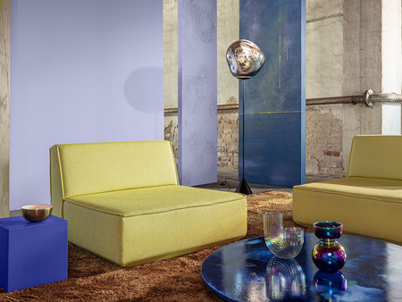 Modern eingerichtete Lounge in industriellem Raum mit gelben Sesseln, braunem Teppich, blauem Couchtisch und Glasdeko vor lila und blauen Wandpaneelen.